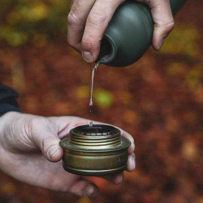 Hand pouring liquid fuel from an olive BRS aluminum bottle into a brass camping stove burner outdoors