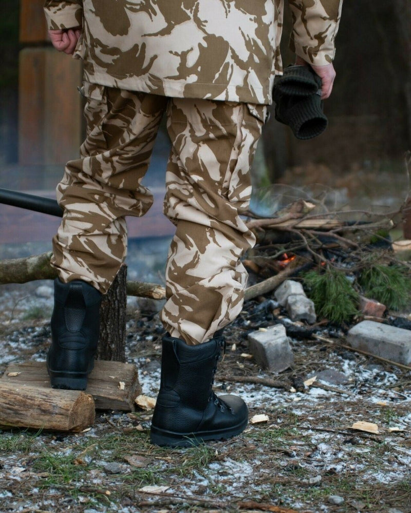 Rear view of person wearing desert camouflage British army combat trousers and black boots near a campfire outdoors